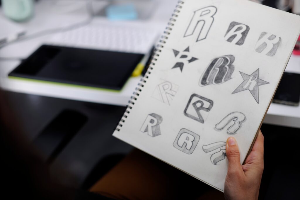 Hand holding a sketchbook displaying various logo design concepts, including stylized letters and shapes, emphasizing creative branding ideas for businesses.